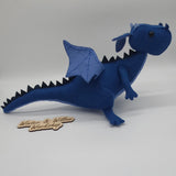 Handmade blue felt dragon plush with black button eyes, dark felt back spikes, and a stitched light blue belly. The soft dragon stands with a curved tail and small wings.