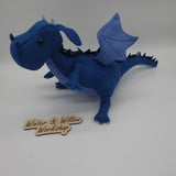 Handmade blue felt dragon plush with black button eyes, dark felt back spikes, and a stitched light blue belly. The soft dragon stands with a curved tail and small wings.