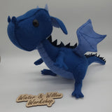Handmade blue felt dragon plush with black button eyes, dark felt back spikes, and a stitched light blue belly. The soft dragon stands with a curved tail and small wings.