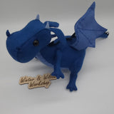 Handmade blue felt dragon plush with black button eyes, dark felt back spikes, and a stitched light blue belly. The soft dragon stands with a curved tail and small wings.