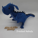 Handmade blue felt dragon plush with black button eyes, dark felt back spikes, and a stitched light blue belly. The soft dragon stands with a curved tail and small wings.