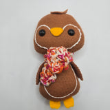 Brown plush gingerbread penguin with a multicolored red, orange, pink, and white scarf on a white background