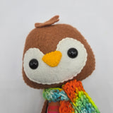 Close-up of a plush Gingerbread Penguin face with rainbow scarf on a light gray background