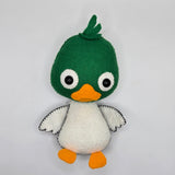 Handmade felt duck plush with a green head, white body, small white wings with black stitching, and bright orange beak and feet. The soft stuffed duck features large round eyes and a small tuft of felt feathers on top of its head.