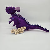 Handmade purple felt dragon plush with a light lavender belly, small white horns, and black button eyes. The dragon is decorated with shimmering sequins across its head, wings, and body.