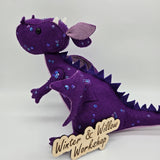 Handmade purple felt dragon plush with a light lavender belly, small white horns, and black button eyes. The dragon is decorated with shimmering sequins across its head, wings, and body.