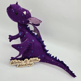 Handmade purple felt dragon plush with a light lavender belly, small white horns, and black button eyes. The dragon is decorated with shimmering sequins across its head, wings, and body.