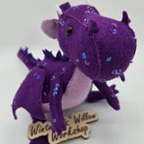 Handmade purple felt dragon plush with a light lavender belly, small white horns, and black button eyes. The dragon is decorated with shimmering sequins across its head, wings, and body.