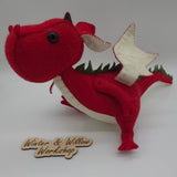 Handmade red felt dragon plush with white wings, small horns, and black button eyes. The soft stuffed dragon stands upright with a curved tail and stitched details.