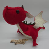 Handmade red felt dragon plush with white wings, small horns, and black button eyes. The soft stuffed dragon stands upright with a curved tail and stitched details.