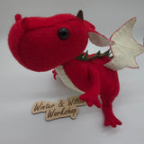 Handmade red felt dragon plush with white wings, small horns, and black button eyes. The soft stuffed dragon stands upright with a curved tail and stitched details.
