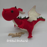 Handmade red felt dragon plush with white wings, small horns, and black button eyes. The soft stuffed dragon stands upright with a curved tail and stitched details.