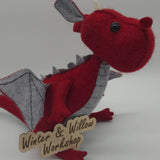 Handmade red felt dragon plush with gray wings, small white horns, and black button eyes. The soft dragon has a stitched gray belly and stands upright with outstretched wings.