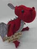 Handmade red felt dragon plush with gray wings, small white horns, and black button eyes. The soft dragon has a stitched gray belly and stands upright with outstretched wings.