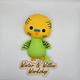 Handmade felt parakeet plush with a bright yellow head, green body, and patterned green wings. The cute bird has black button eyes, a small orange beak with a blue cere, and stitched feather details.