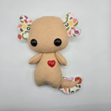 Stuffed axolotl plush resembling a pizza with a pepperoni heart and pizza topping patterned ears and tail on a light gray background