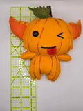 Handmade pumpkin gremlin felt plush with an orange pumpkin body, pointy ears, and stitched pumpkin lines. The character has one black button eye, a winking stitched eye, and a playful tongue sticking out with a small green leaf and stem on top.