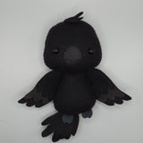 Handmade black raven felt plush with black button eyes, layered felt wings, and a textured beak. The soft stuffed bird features small tail feathers and a tuft of feathers on top of its head, creating a cute raven-inspired plush design.